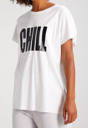 T-shirt blanc ample avec un grand texte noir "CHILL" porté par une personne aux cheveux bouclés, debout devant un fond clair uni.