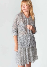 Abito lungo e trasparente con maniche lunghe, decorato con un motivo floreale in grigio e bianco, dotato di una chiusura frontale con bottoni e gonna plissettata. Tessuto leggero e testurizzato.