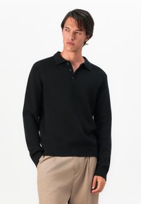 Suéter polo preto de mangas longas, textura canelada, com paletó de três botões, design ajustado, combinado com calças beje claras.