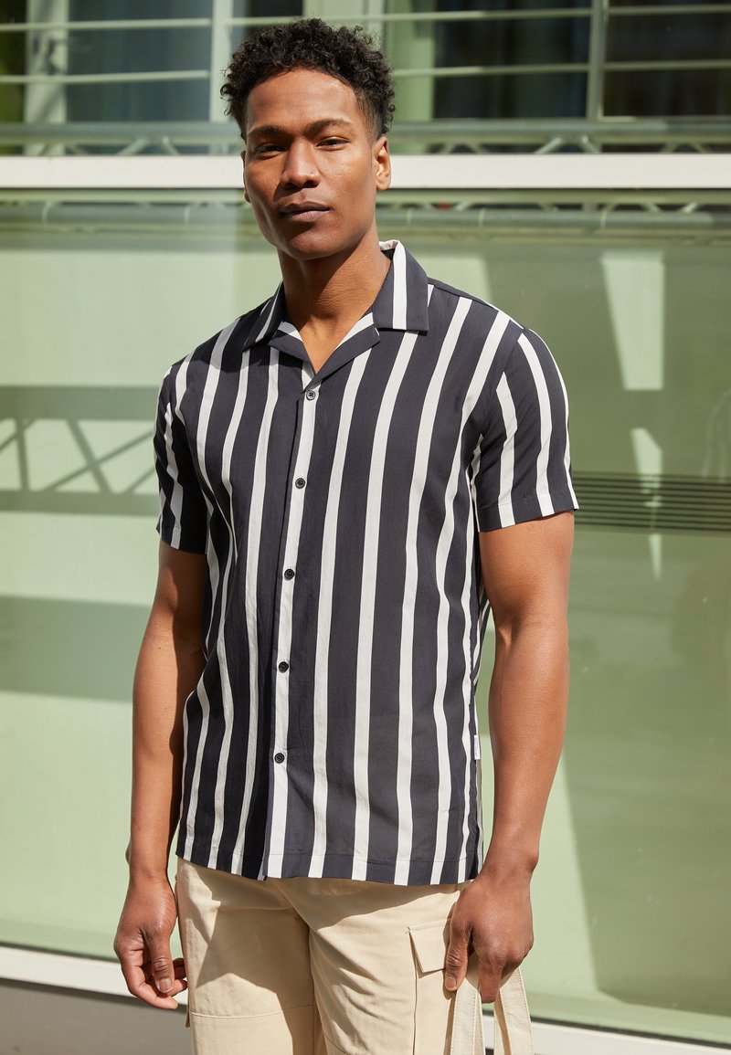 Chemise à manches courtes boutonnée à rayures verticales noires et blanches, avec un col et une coupe décontractée. Texture de tissu lisse.