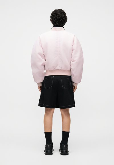 Άτομο με σγουρά μαλλιά στέκεται πλάτη, φοράει ανοιχτό ροζ μπουφάν, μαύρο σορτσάκι με λευκή ραφή, μαύρες κάλτσες και μαύρα παπούτσια.