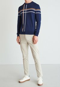 Marineblauer Pullover mit weißen und orangefarbenen Streifenakzenten, Crew-Neck-Design, kombiniert mit hellbeigen Hosen und weißen Sneakers.
