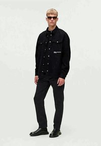 Crna traper jakna s dugmadi na pritiskanje, dva prednja džepa i bijelim otisnutim logotipom. Uparena s crnim hlačama i elegantnim crnim čizmama.