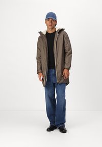 Brown padded jacket with a fleece-lined collar, zip closure, and elastic cuffs, worn over a black sweater and blue jeans. Blue cap.