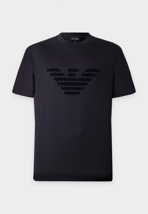 Jeune homme portant un t-shirt graphique noir, un jean gris et des baskets noires, debout devant un fond clair uni.