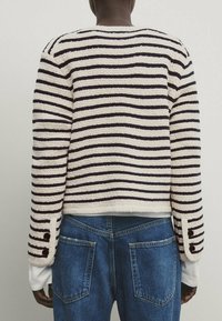 Person wearing a cream sweater with dark horizontal stripes and two buttons on each cuff, paired with blue denim jeans seen from the back.
