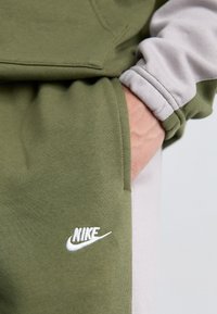 Közelkép egy személy kezéről, amely zöld és bézs sportöltözet zsebében van, fehér Nike logóval az anyagon.