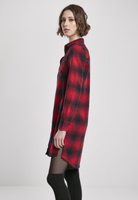 Robe chemise à carreaux rouge et noir avec un col, des manches longues et des fentes sur les côtés. Tissu texturé avec une coupe ample, associée à des chaussettes noires jusqu'aux genoux.