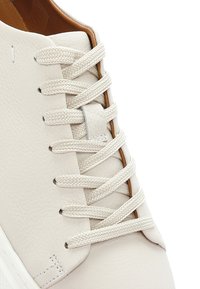 Witte leren sneaker met gestructureerde veterlinten in een kruislingse patroon, voorzien van een lichtbruine voering en versterkte stiksels nabij de zool.