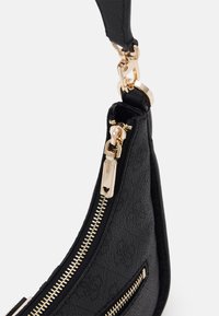 Sac à main noir texturé avec deux fermetures éclair dorées et une boucle dorée fixant la sangle noire au corps du sac sur un fond blanc.