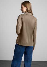 Pitkähihaista ruskeaa puseroa, jossa on kaulus, yhdistettynä löysiin sinisiin farkkuihin. Kangas näyttää pehmeältä ja sileältä yksinkertaisella muotoilulla.