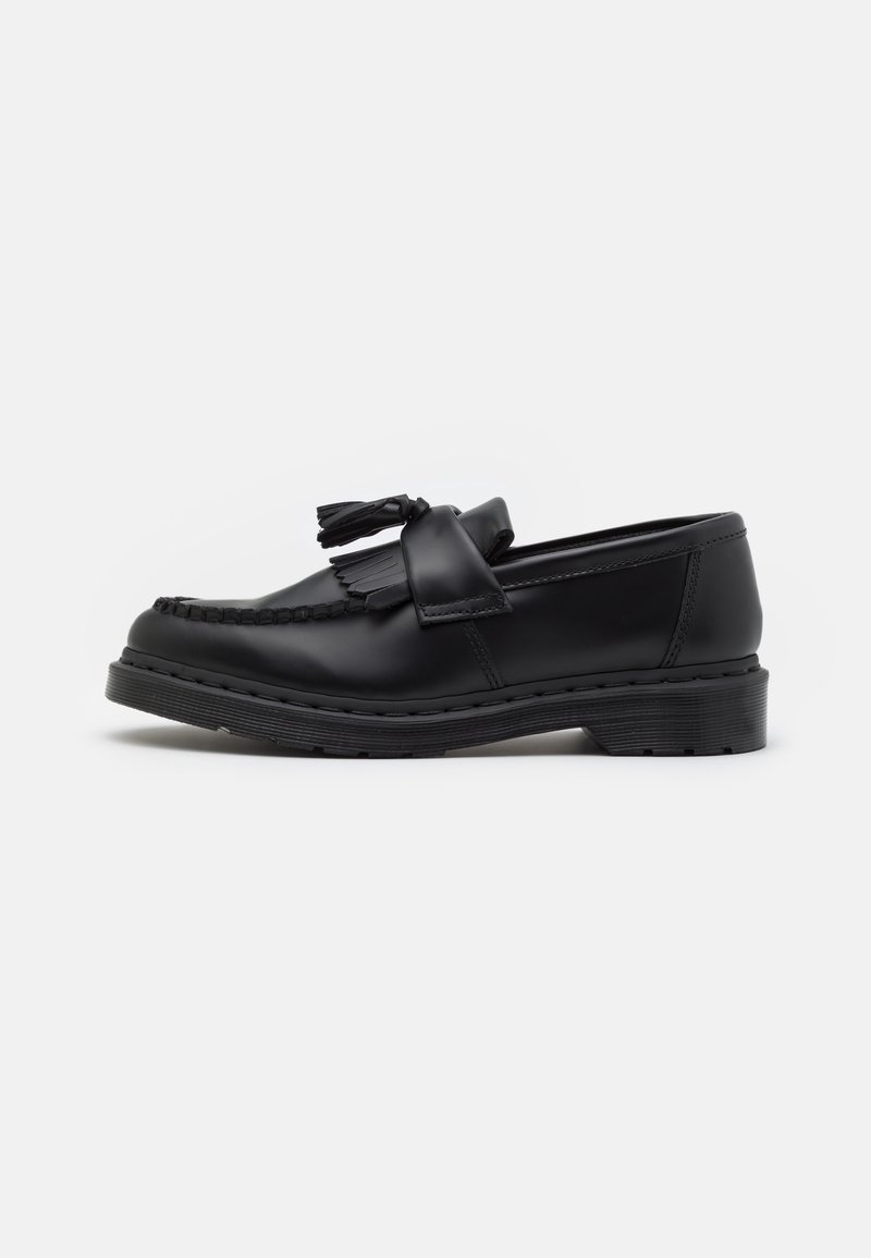Mocassin en cuir noir avec pompons, détail de franges et semelle épaisse cousue, présenté de profil sur un fond uni.