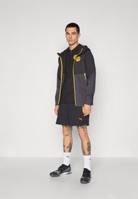 Sort og grå lynlåstrøje med gule detaljer, parret med en sort polo og mørke shorts. Sorte atletisk sko fuldender outfittet.