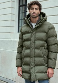Olivengrøn pufferjakke med hætte, der har en firkantet quiltet design, lynlåslukning og forlommer. Båret over et gråt lag.