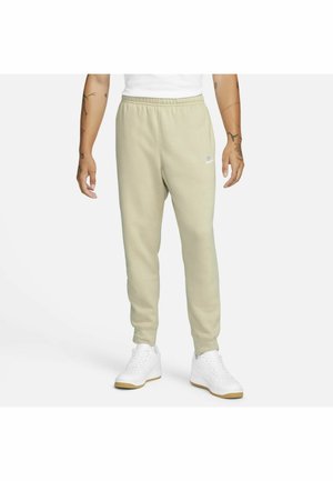 Homme portant un pantalon de jogging Nike beige clair et des baskets blanches, debout avec les bras détendus devant un fond blanc uni.