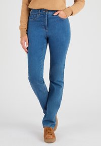 Vaqueros de denim azul con corte ajustado, que cuentan con cinco bolsillos, trabillas para cinturón y costuras sutiles. Combinados con zapatos de ante marrones.