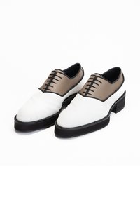 Witte leren schoenen met een bruine leren bovenkant, zwarte veters en een getextureerd ontwerp, met een dikke zwarte zool en een ronde neus.