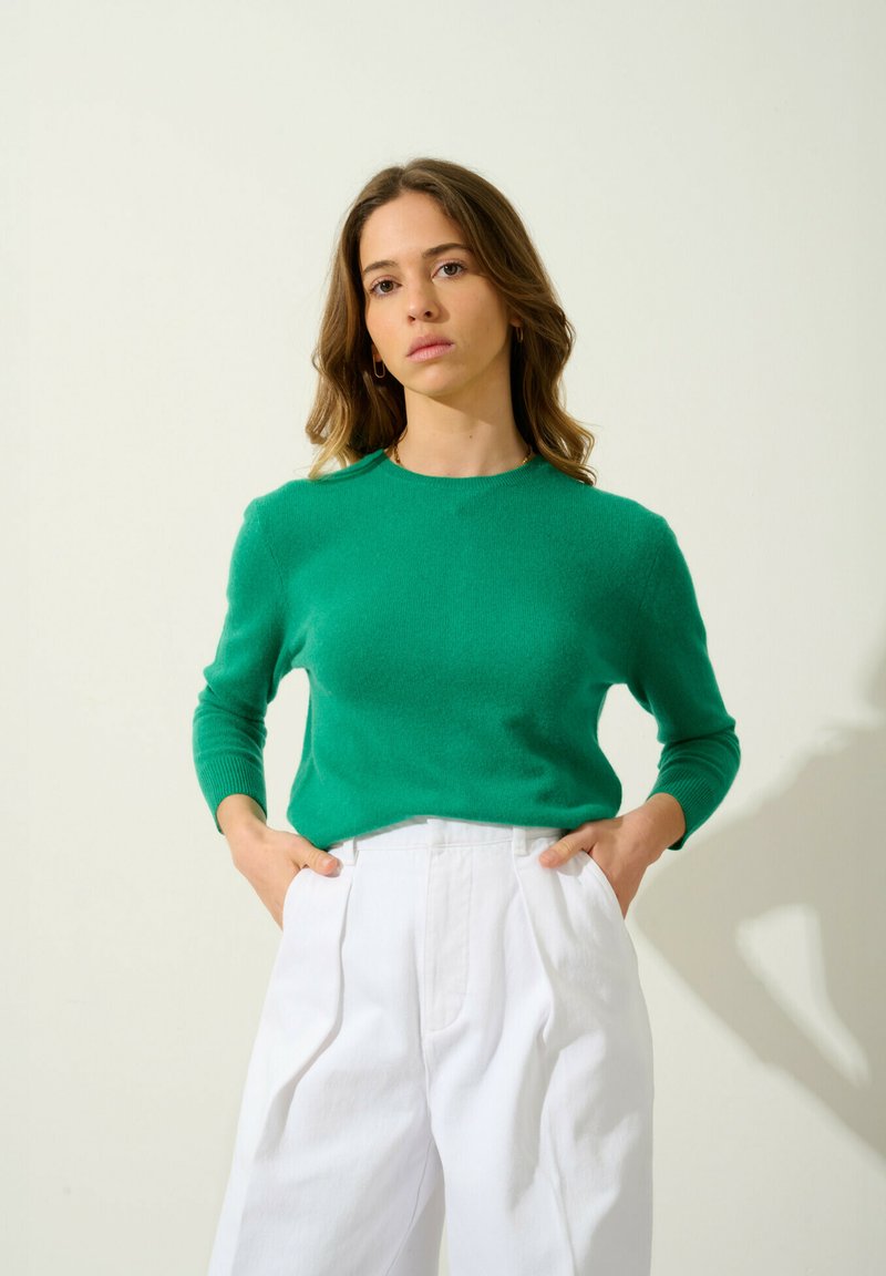 Zielony sweter dzianinowy z długimi rękawami, okrągłym dekoltem i miękką fakturą, w połączeniu z białymi plisowanymi spodniami z szerokimi nogawkami.