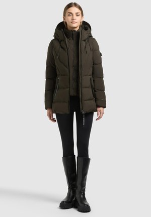 Femme portant une veste matelassée à capuche vert foncé, des leggings noirs et des bottes noires montantes, debout sur un fond clair uni.
