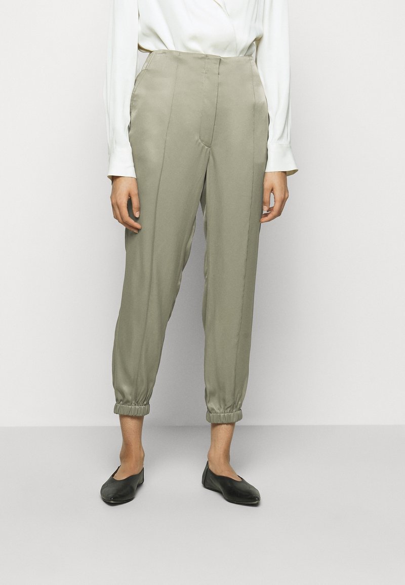Femme portant un pantalon taille haute, fuselé couleur olive clair avec des poignets élastiques, associé à des ballerines noires et un chemisier blanc à manches longues.