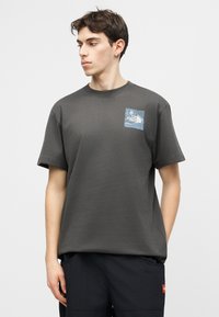 Jeune homme portant un t-shirt gris foncé avec un petit motif bleu et blanc sur la poitrine, debout les mains dans les poches d'un pantalon noir.