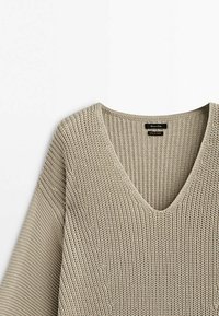Light beige knit sweater with a V-neck and wide, loose sleeves. Features a textured weave and tag with "Made in Sydney."