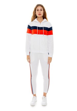 Une femme porte un survêtement blanc à rayures bleu marine et rouges ainsi que des baskets blanches, sur un fond uni.