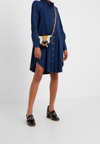 Džínová košilová šaty s knoflíky, dlouhými rukávy a zakřiveným lemem. Černé lodičky na podpatku s kulatým kováním; malá barevná kabelka.