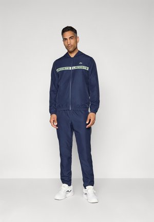 Tuta navy con il branding "LACOSTE" in bianco e verde. Include una giacca con zip, polsini elasticizzati e pantaloni stile jogger. Scarpe da ginnastica bianche.
