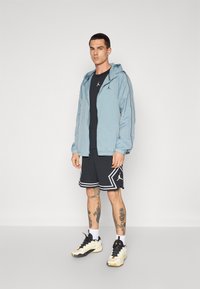 Lysblå vindjakke med hætte, sort skjorte, sorte shorts med hvide detaljer, og sorte sneakers; tatoverede ben synlige.