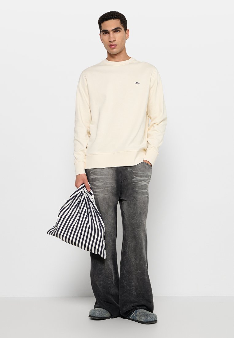 Junger Mann in cremefarbenem Sweatshirt und verblassten schwarzen weiten Jeans, der eine schwarz-weiße gestreifte Tasche hält und vor einem weißen Hintergrund steht.