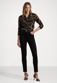 Camicia nera con motivo a catena dorata, jeans neri aderenti e décolleté neri con cinturini. La camicia presenta un colletto e maniche lunghe.