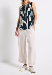 Kvinna iförd ärmlös mönstrad blus, vida krämfärgade byxor och vita sneakers, stående mot en enfärgad vit bakgrund.