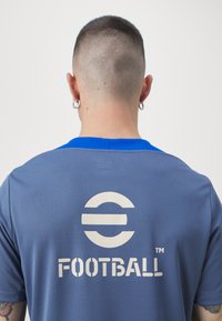 Camiseta atlética azul con una superficie texturizada, que presenta un gran logo circular y la palabra "FÚTBOL" en blanco en la parte posterior.