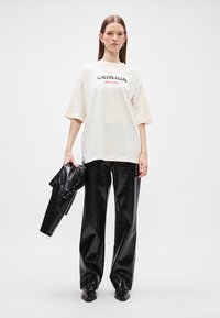 Εκτός λευκό oversized T-shirt με το κείμενο "Calvin Klein New York"; συνδυασμένο με γυαλιστερούς μαύρους παντελόνες από κροκό και μια ταιριαστή τσάντα.