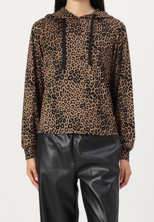 Kobieta w brązowo-czarnym kapturze w cętki lamparta z czarnymi sznureczkami i czarnymi skórzanymi spodniami, stojąca na jednolitym tle.