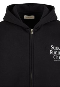 Felpa con zip nera realizzata in misto cotone, dotata di un ampio cappuccio e tasca anteriore. La scritta "Sundays Running Club" è stampata in bianco sul lato sinistro.