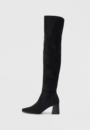 Botte noire en daim montée jusqu'au genou, avec un talon bloc, un bout pointu et un design lisse et sans couture sur un fond clair.