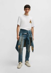 Weißes T-Shirt mit Grafikdruck und kurzen Ärmeln, kombiniert mit verwaschenen blauen Jeans und einer Denimjacke, die um die Taille gebunden ist. Beigefarbene Sneaker.