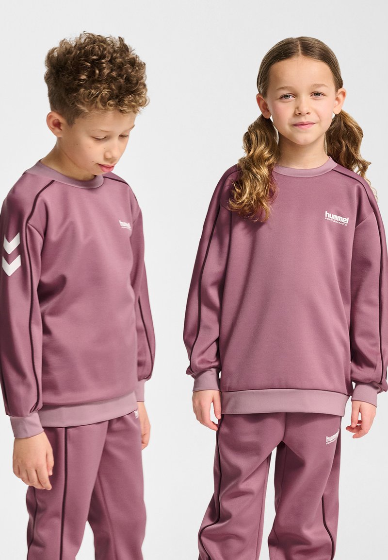 Twee kinderen dragen bijpassende paarse sweaters en broeken van gladde stof. De sweaters hebben contrasterende schouderpanelen en branding.