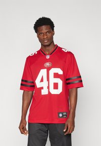 Sarkanais NFL krekls ar melnām svītrām uz piedurknēm, skaitlis "46" baltā krāsā, komandas logo uz krūts, izgatavots no elpojoša materiāla. Zem tā redzami melni šorti.