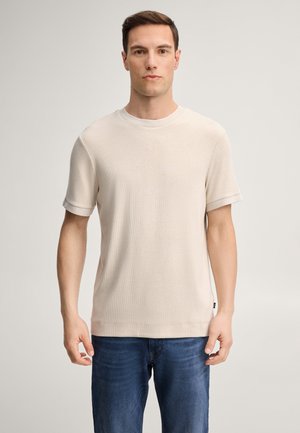 Man met kort bruin haar staat recht vooruit, draagt een beige shirt met korte mouwen en blauwe spijkerbroek tegen een effen lichte achtergrond.
