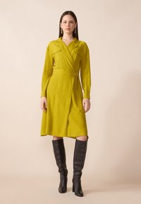 Långärmad omlotklänning i livlig limegrön texturerad tyg, med b�älte i midjan och spetsig krage, kombinerad med knähöga svarta stövlar.