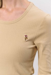 Beige ribbad långärmad topp med rund halsringning och en liten broderad blomsterdesign på vänstra bröstet. Mjuk tygstruktur.