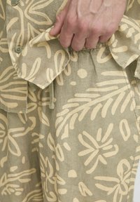 Chemise en lin avec un motif floral beige sur une base verte, dotée d'une fermeture à boutons et d'une main tenant le tissu.
