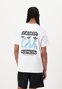 White cotton t-shirt featuring large black text and blue floral graphics on the back, with "ADIDAS ORIGINALS" and tagline at the bottom.