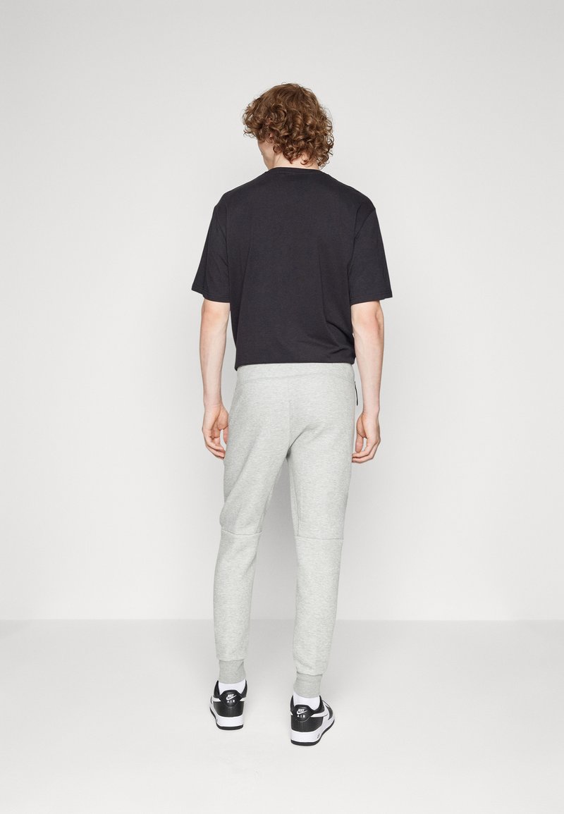 Schwarzes T-Shirt mit kurzen Ärmeln kombiniert mit hellgrauen, tapered Jogginghosen, die über gerippte Bündchen und einen Kordelzugbund verfügen. Schwarze Sneakers.