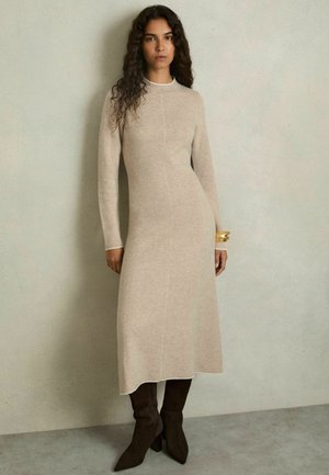 Beiges Strickkleid mit langen Ärmeln und einem hohen Kragen, mit vertikalen Nahttdetails. Kombiniert mit braunen Wildleder-Stiefeletten.