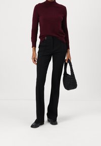 Pull bordeaux, ajusté, texture côtelée. Pantalon noir sur mesure avec des coutures latérales. Sac à main tissé noir avec une courte anse. Chaussures noires.