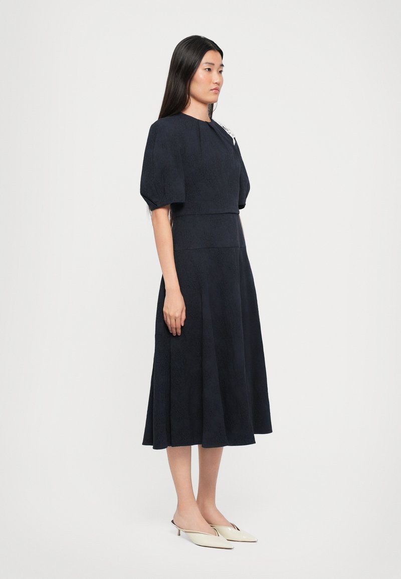 Navyblauwe jurk met korte, gezwollen mouwen en een aansluitend lijfje. Glad textuur, A-lijn silhouet en subtiel patroon door de hele jurk.